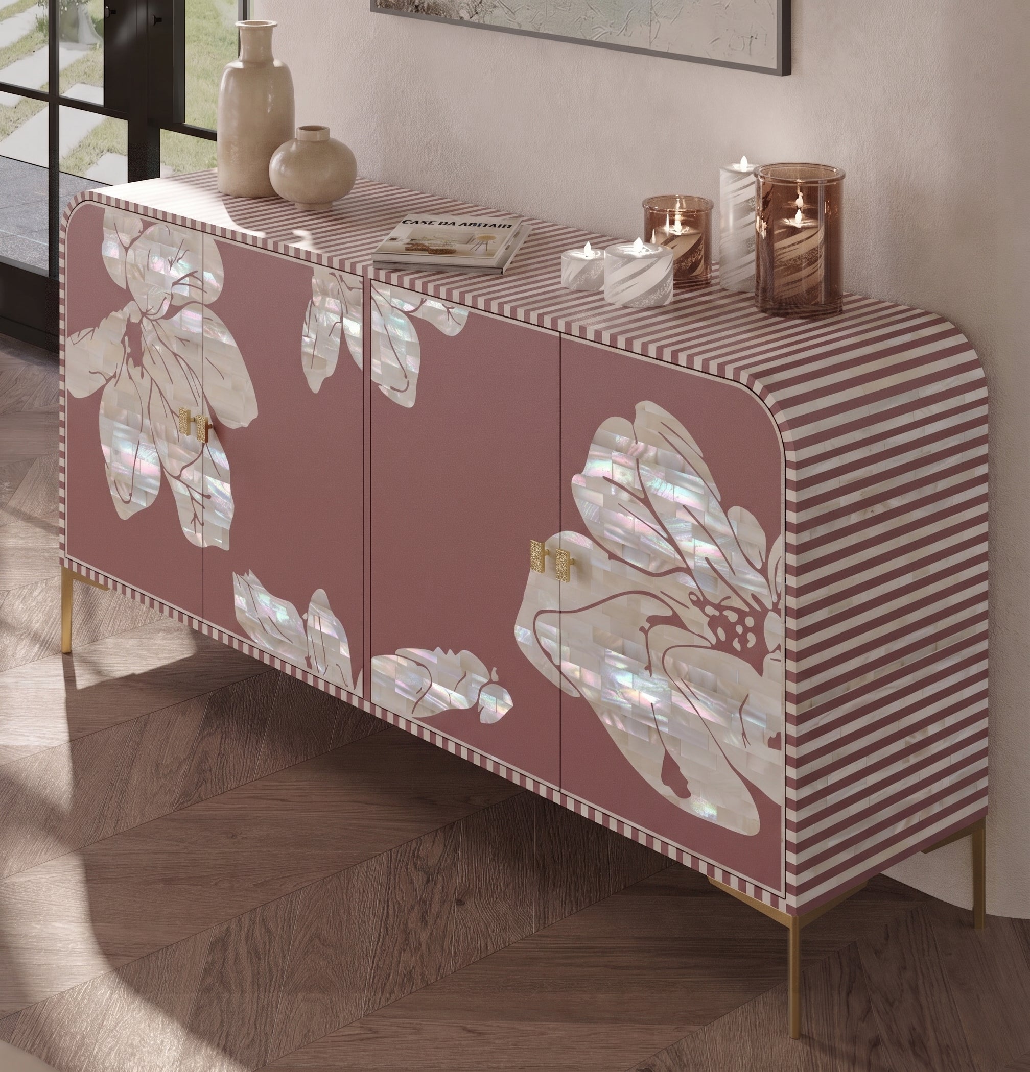 A modern sideboard cabinet with a dusty rose pink finish, featuring large iridescent mother-of-pearl floral motifs on the doors and a pink-and-white striped top panel. The cabinet is styled with decorative vases and candles on a wooden herringbone floor.