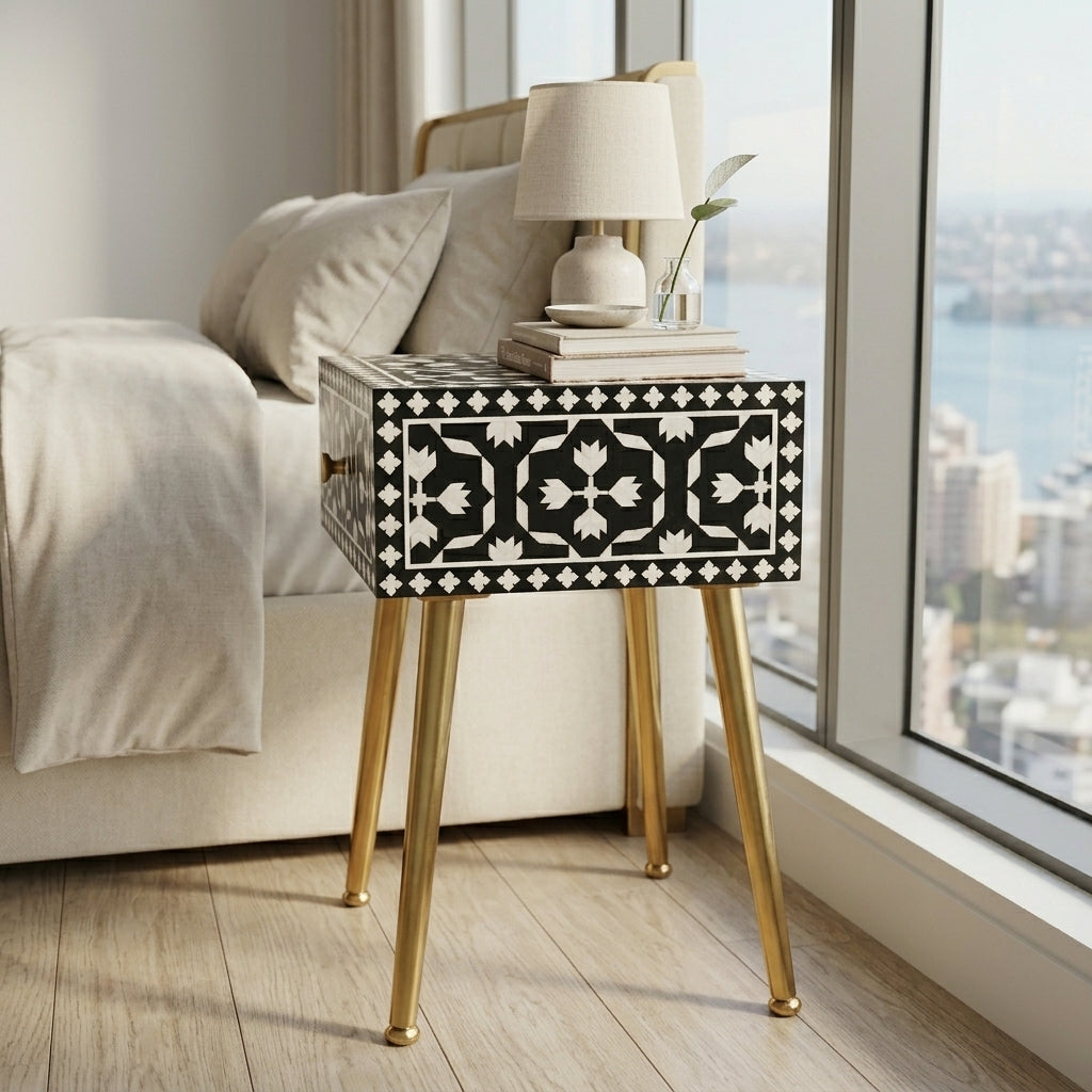 A rectangular bedside table with a black and white geometric inlay pattern on the front and gold-tone metal legs, styled next to a bed in a bright bedroom.