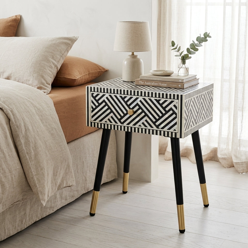 A modern bedside table with a black and white geometric mother of pearl inlay pattern, black legs with gold metal caps, styled next to a bed in a bright bedroom.