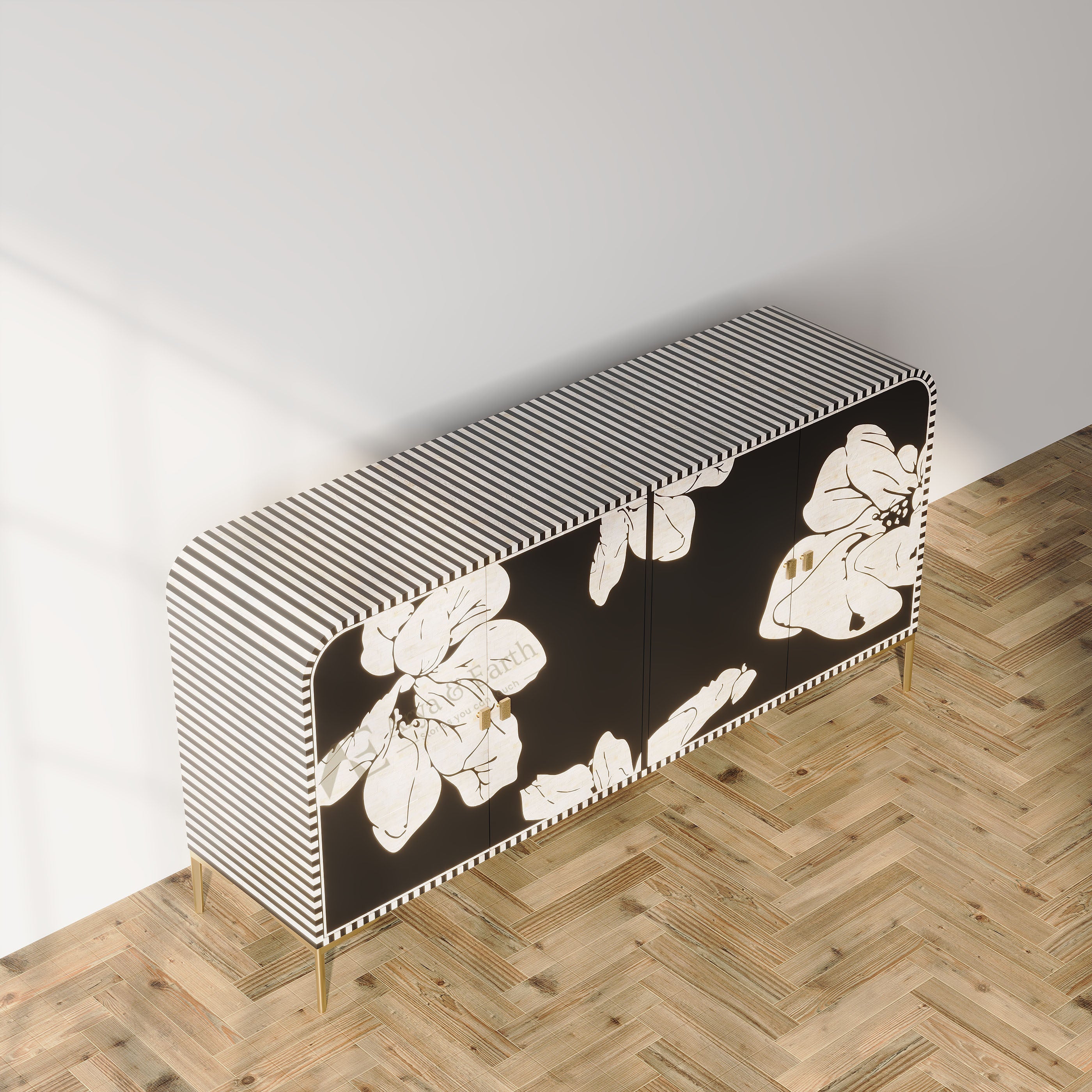 A modern sideboard or credenza with a black and white striped top and sides, black front doors with large white floral motifs, and slim gold legs, shown in a styled room setting with a light wood herringbone floor and white wall.