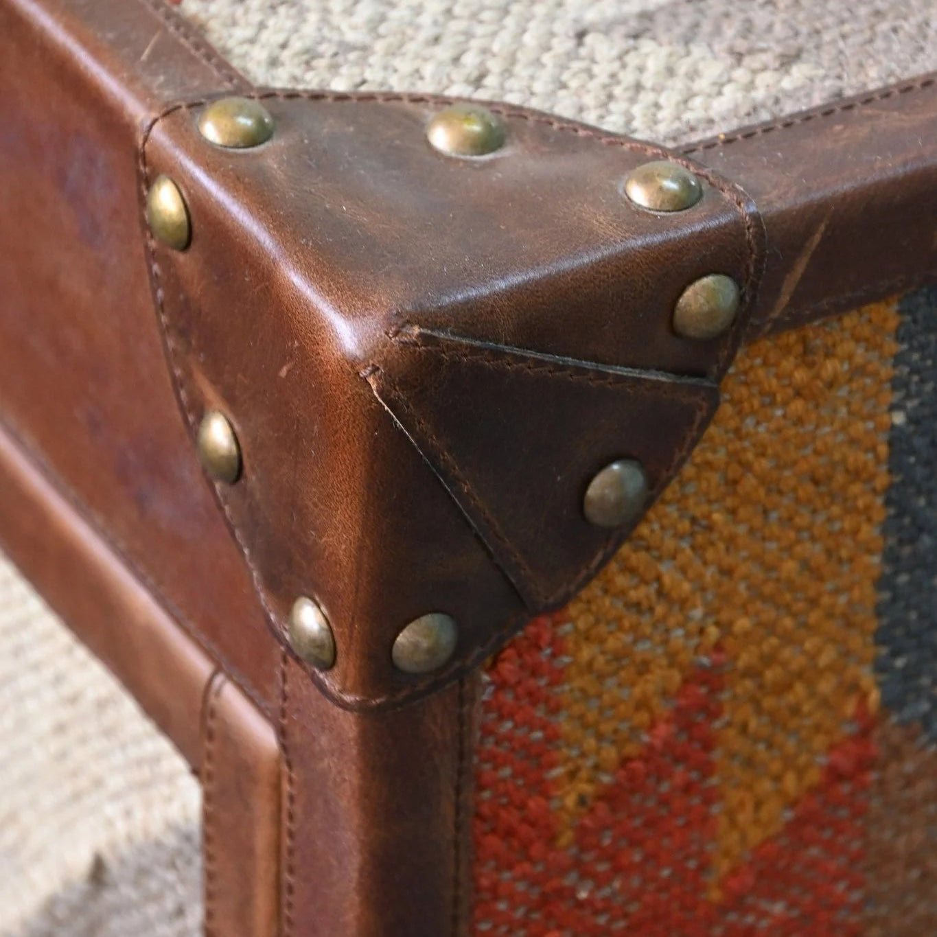 Close-up of a brown leather suitcase corner with brass studs on a textured fabric background.