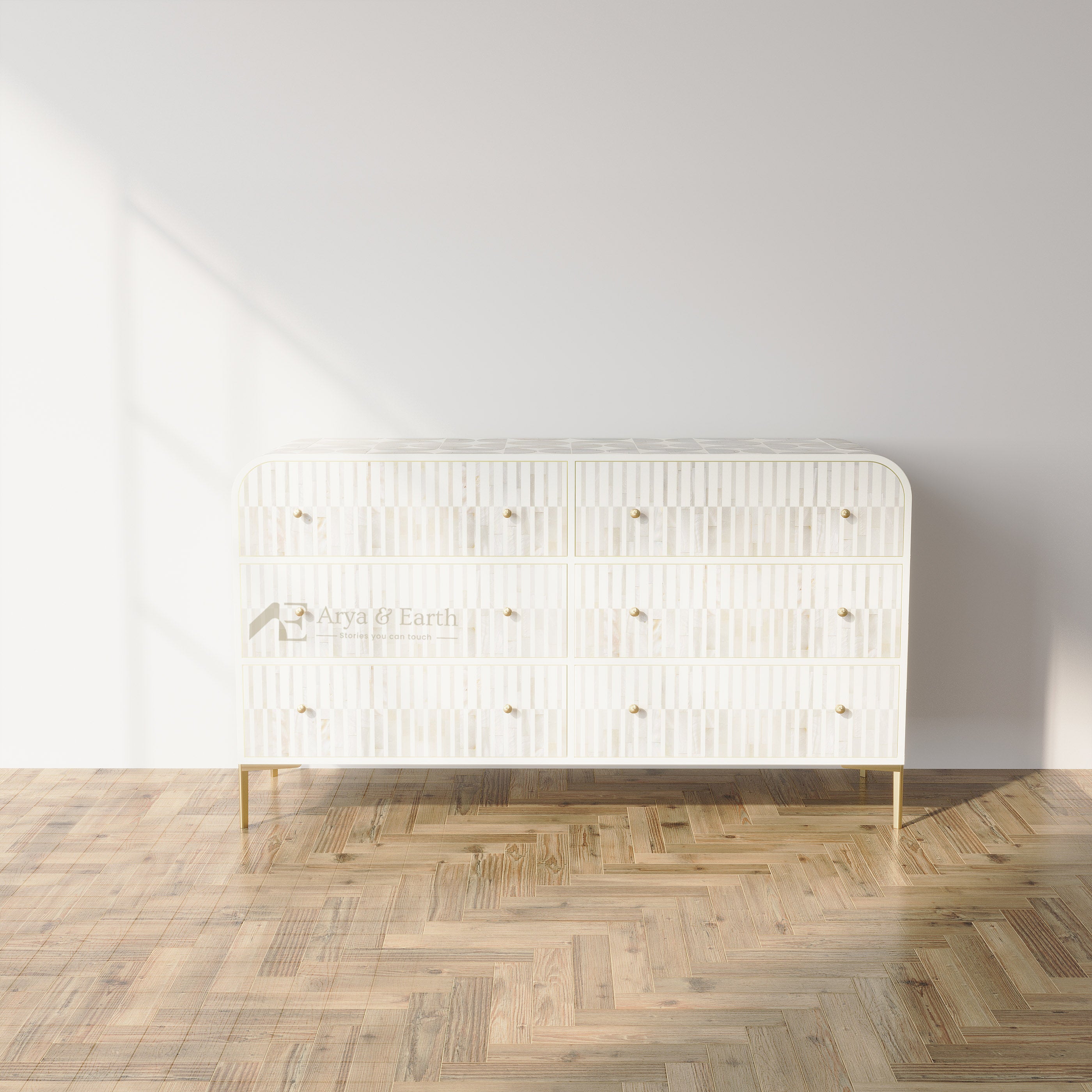 A modern white three-drawer dresser with vertical ribbed detailing and small gold knobs, standing on slim gold legs in a room with a light gray wall and herringbone wood floor.