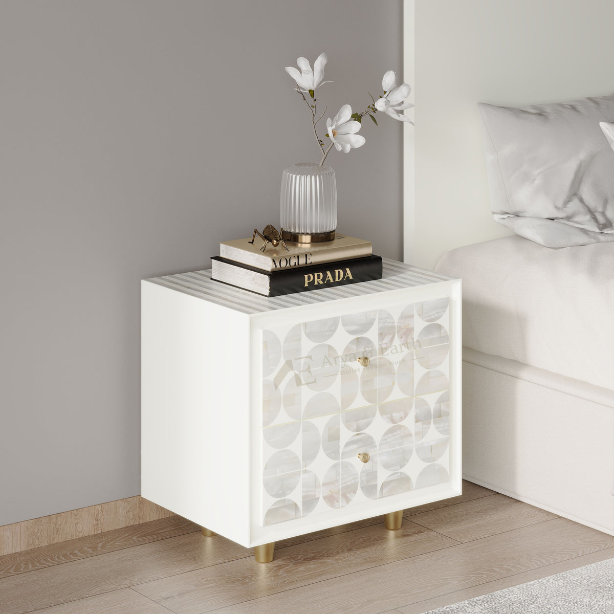 A white bedside cabinet with a geometric patterned glass front and gold legs, styled next to a bed with books and a vase on top.