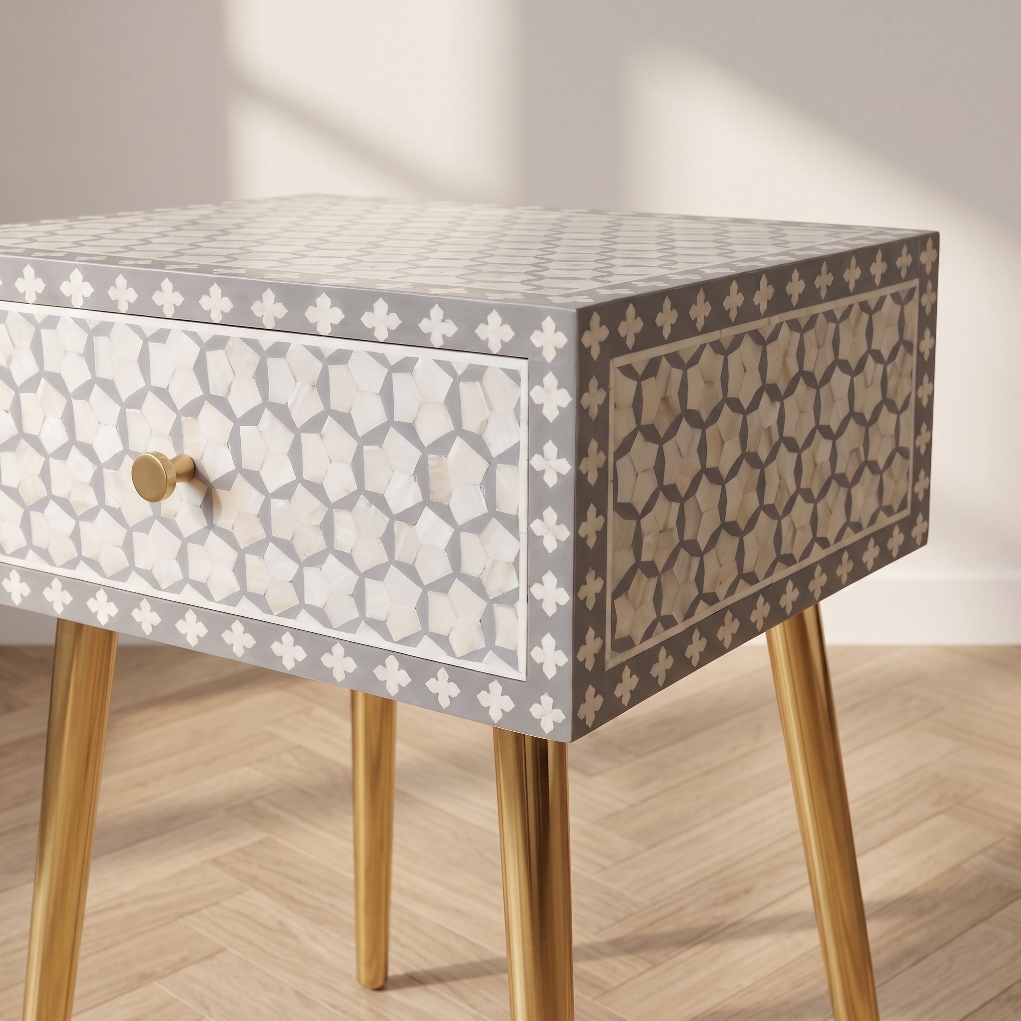 A modern side table with a geometric bone-inlay pattern in beige, brown, and white, featuring tapered gold legs and a single drawer with a brass knob, set against a neutral wall and light wood floor.