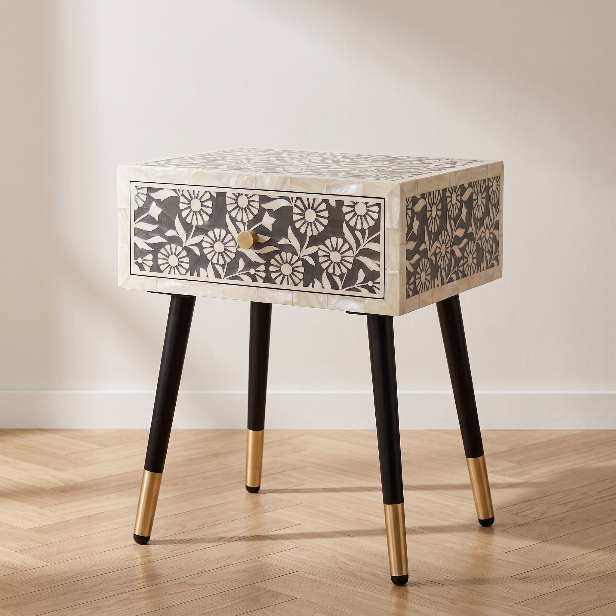 A small rectangular side table with an inlay patterned top and drawer front, black legs, and gold-toned metal foot caps, shown in a room with a light wood floor and off-white wall.