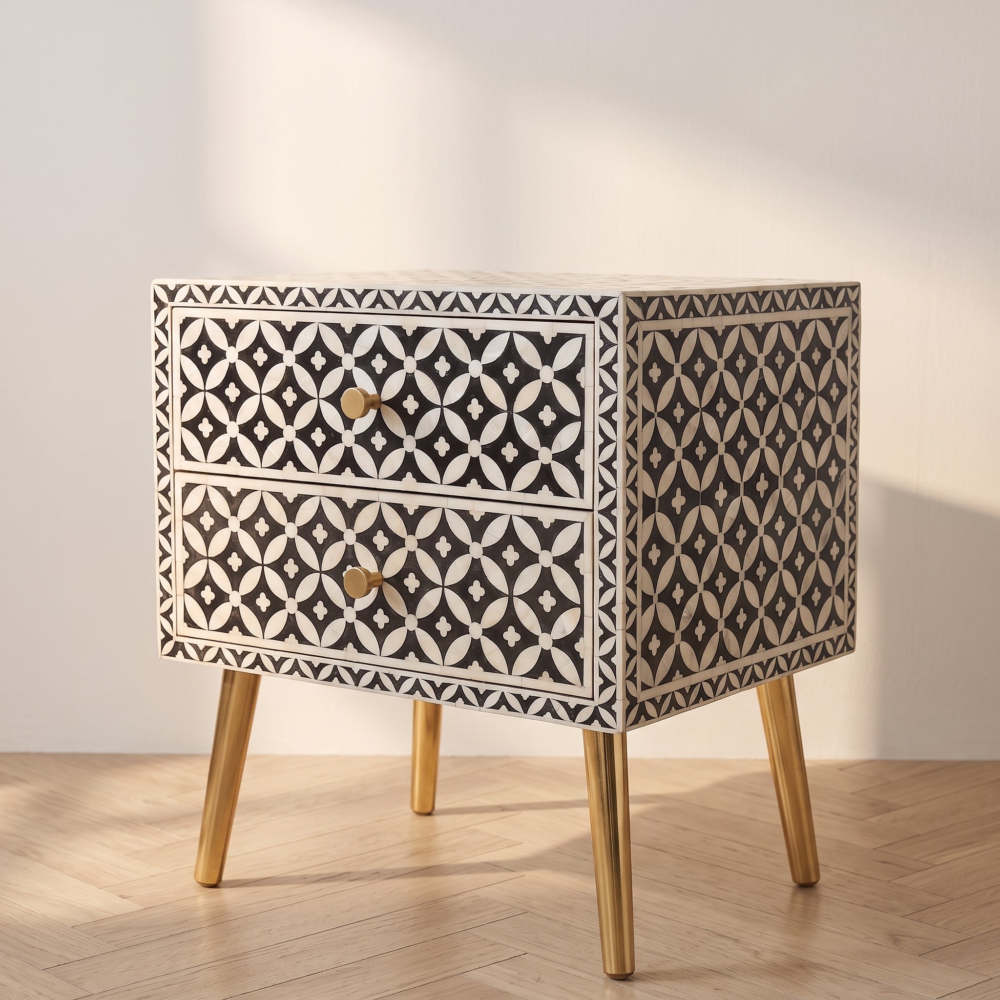 A two-drawer nightstand with a black and white geometric bone-inlay pattern and light wood legs, shown on a light wood herringbone floor against a neutral wall with soft shadows.
