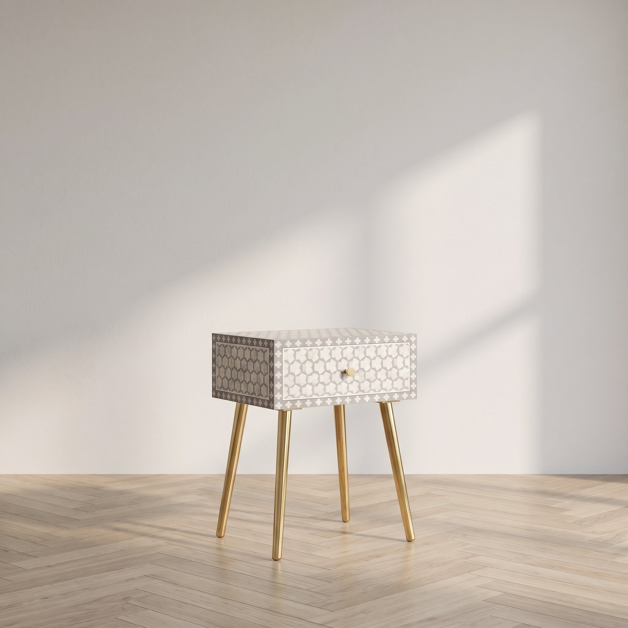A small modern side table with a patterned cream and gray drawer front, gold-tone legs, and a single round gold pull, shown in a minimalist room with a light wood floor and wall shadow.