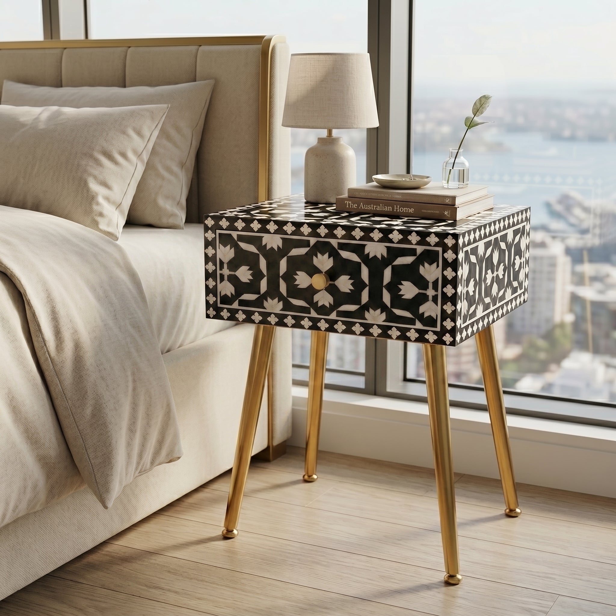 A black and beige bone-inlay nightstand with gold metal legs sits beside a bed in a modern bedroom with a large window overlooking a city and water.