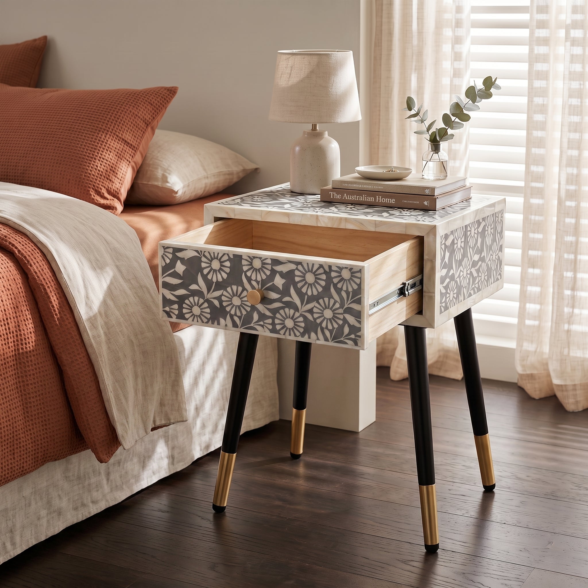 A modern bedside table with a floral-patterned drawer front, white block base, black tapered legs with gold metal caps, and an open drawer showing light wood interiors. It is styled in a bedroom next to a bed with rust-orange bedding, with a lamp and books on top.