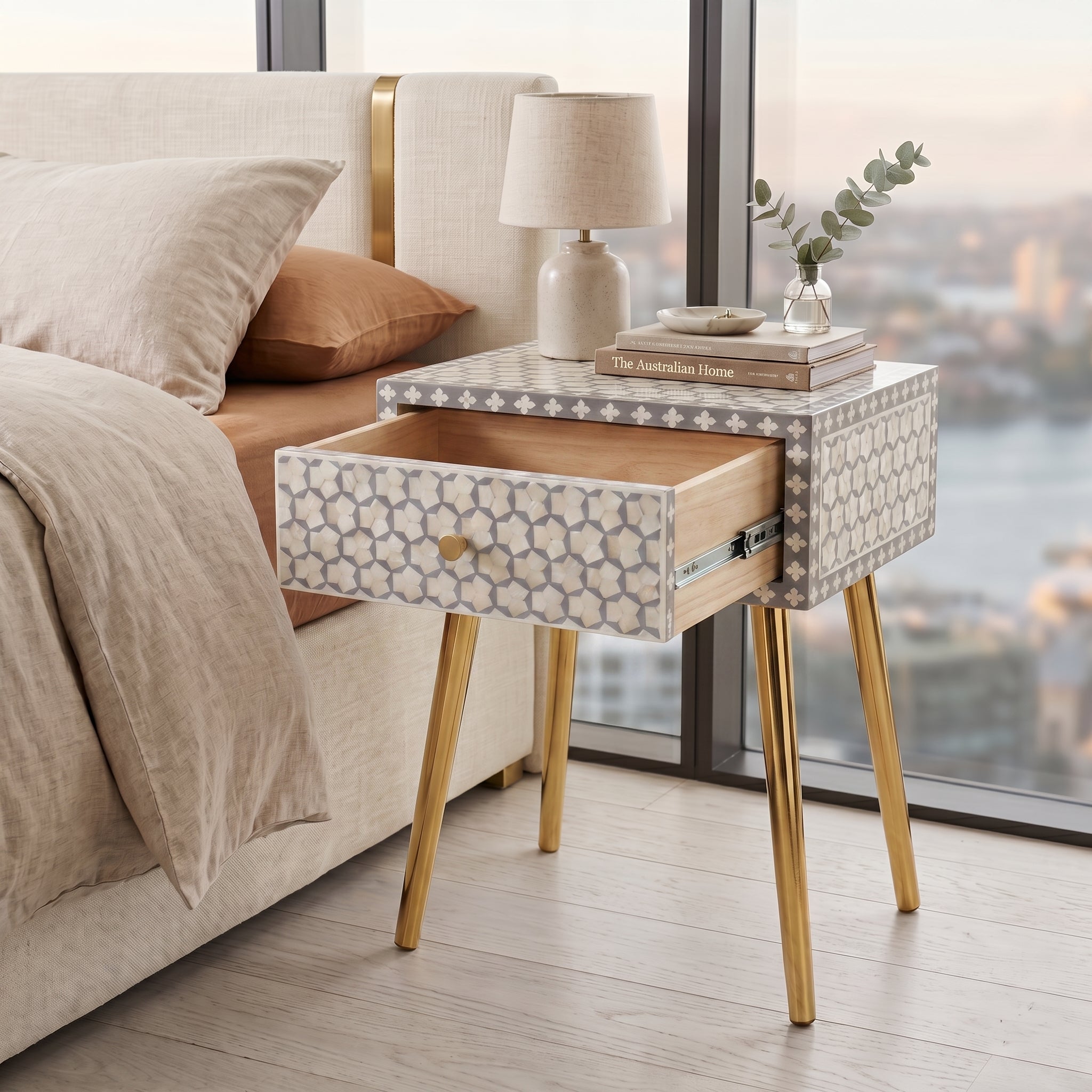 A modern bedside table with a patterned cream and gray drawer front, gold-tone legs, and an open drawer, styled next to a bed in a bedroom with a city view outside.