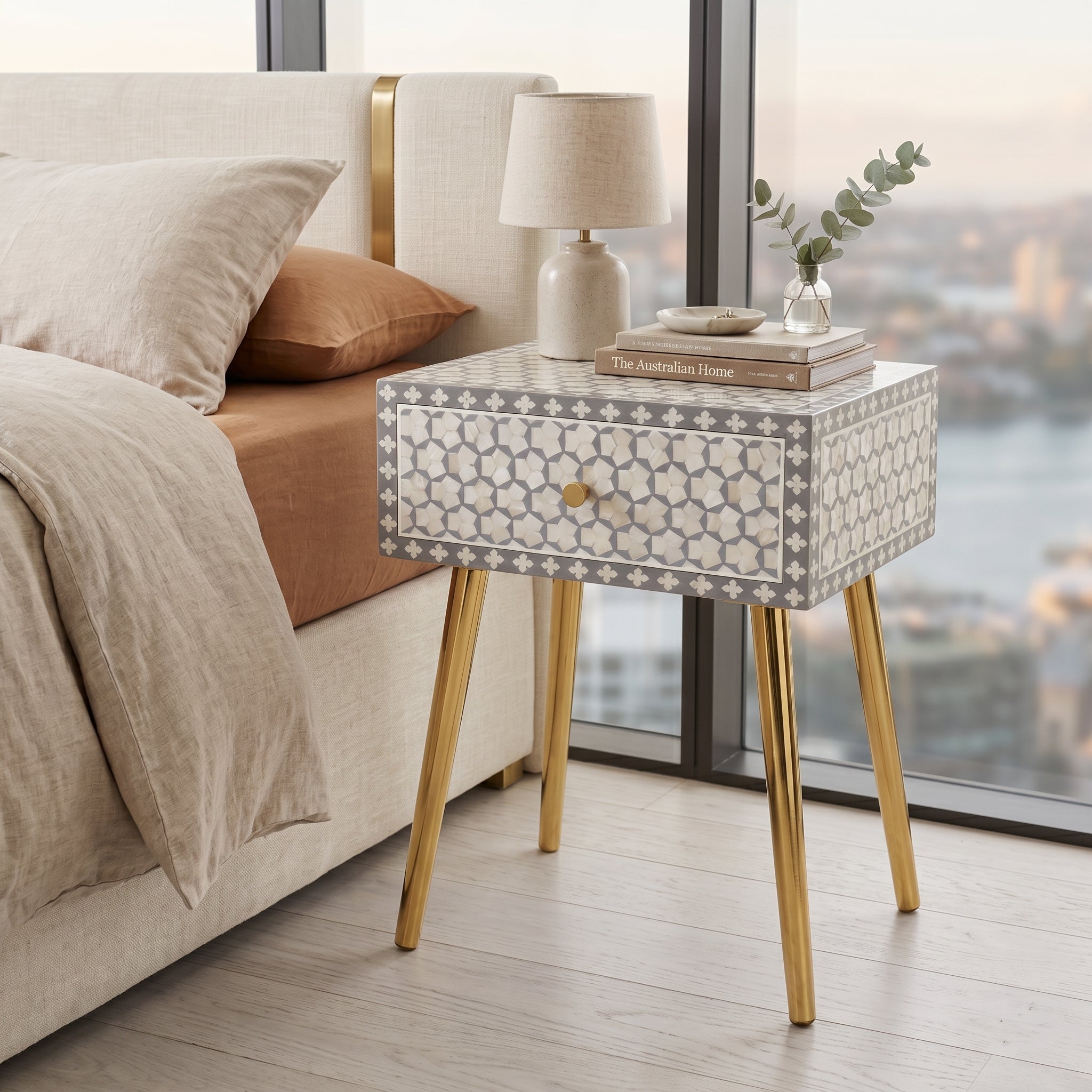 A modern bedside table with a white and gray geometric bone-inlay pattern and gold legs, styled next to a bed in a bright bedroom with a city view outside.