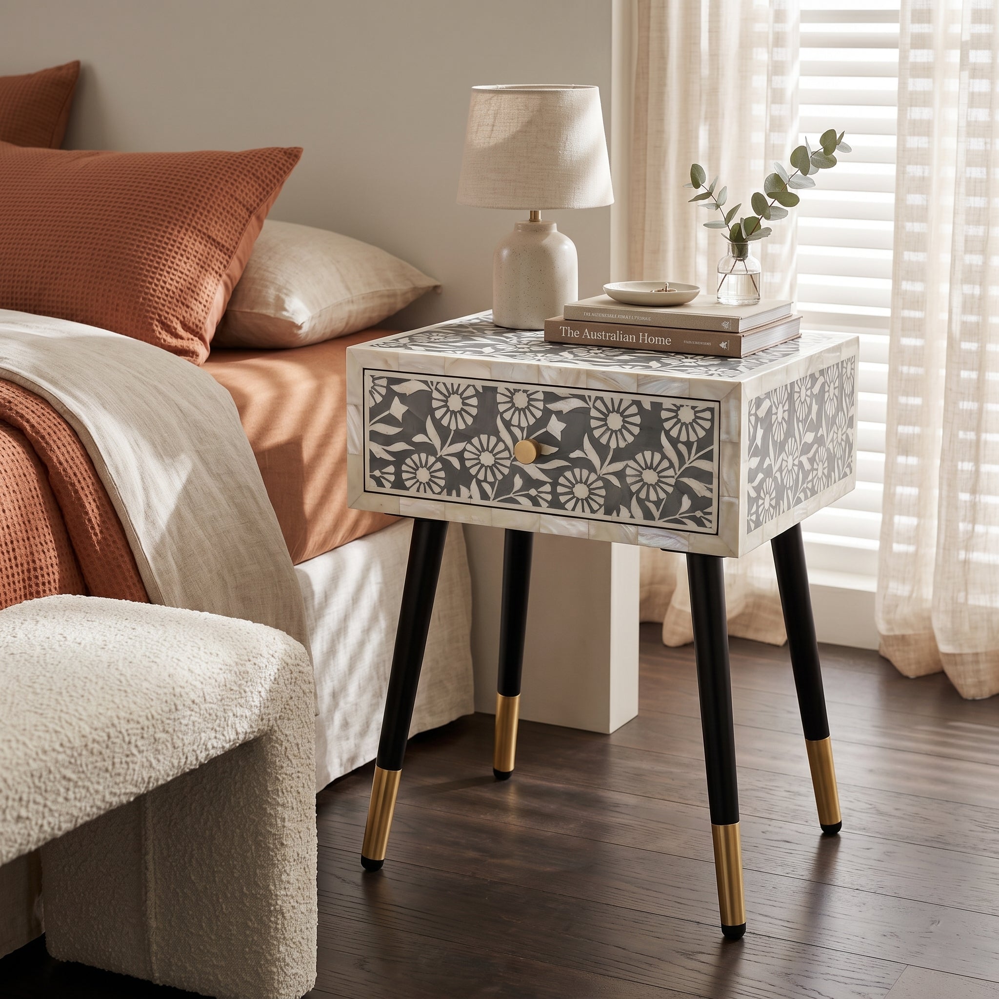 A single-drawer nightstand with a white and gray floral bone-inlay pattern, black legs, and gold metal tips, styled next to a bed in a bedroom.