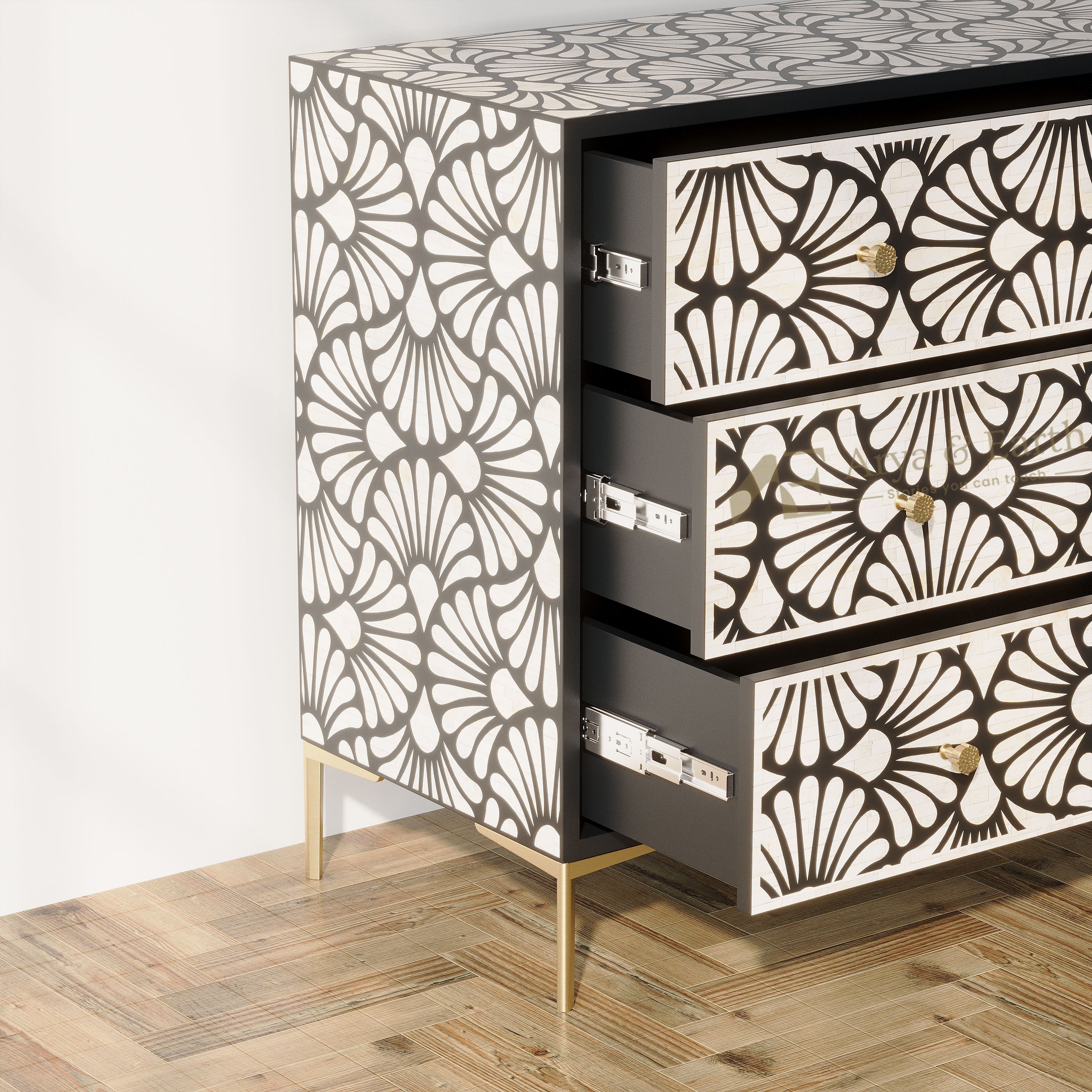 A three-drawer chest of drawers with a black and white floral fan pattern on the exterior and interior drawer fronts, supported by slim gold-toned legs. Two drawers are open, showing a dark gray interior, set against a plain white wall and a light wood parquet floor.