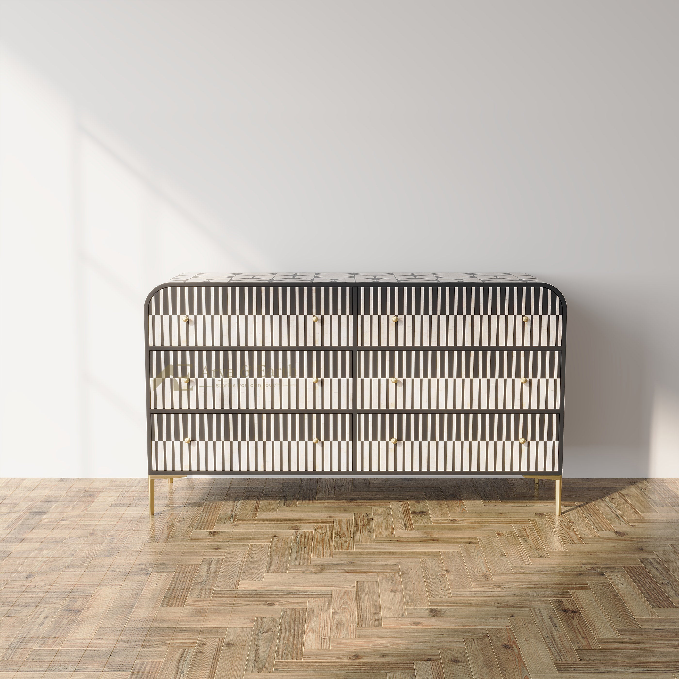 A modern three-drawer chest of drawers with a black frame, white inlay detailing, and slim gold legs, set against a minimalist white wall and light wood herringbone parquet floor.