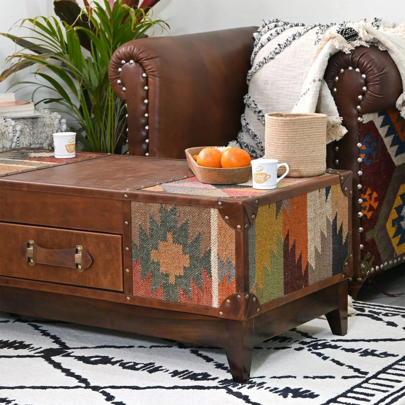 Brown leather sofa with a patterned coffee table, oranges, and a cup on a white wall background.