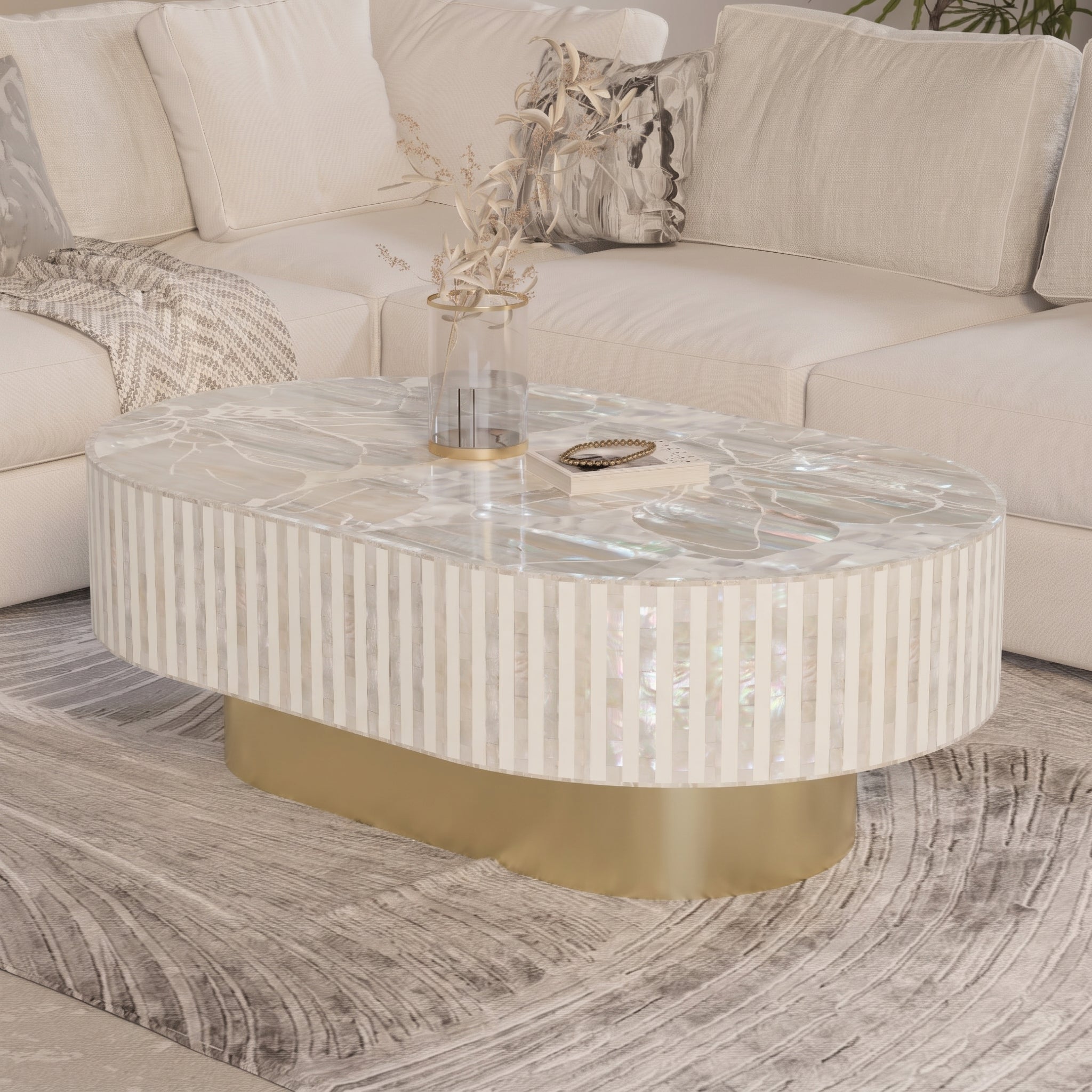 An oval coffee table with a white and iridescent striped shell-look body, a clear glass vase on top, and a brushed gold metal base, set in a modern living room next to a cream sofa.