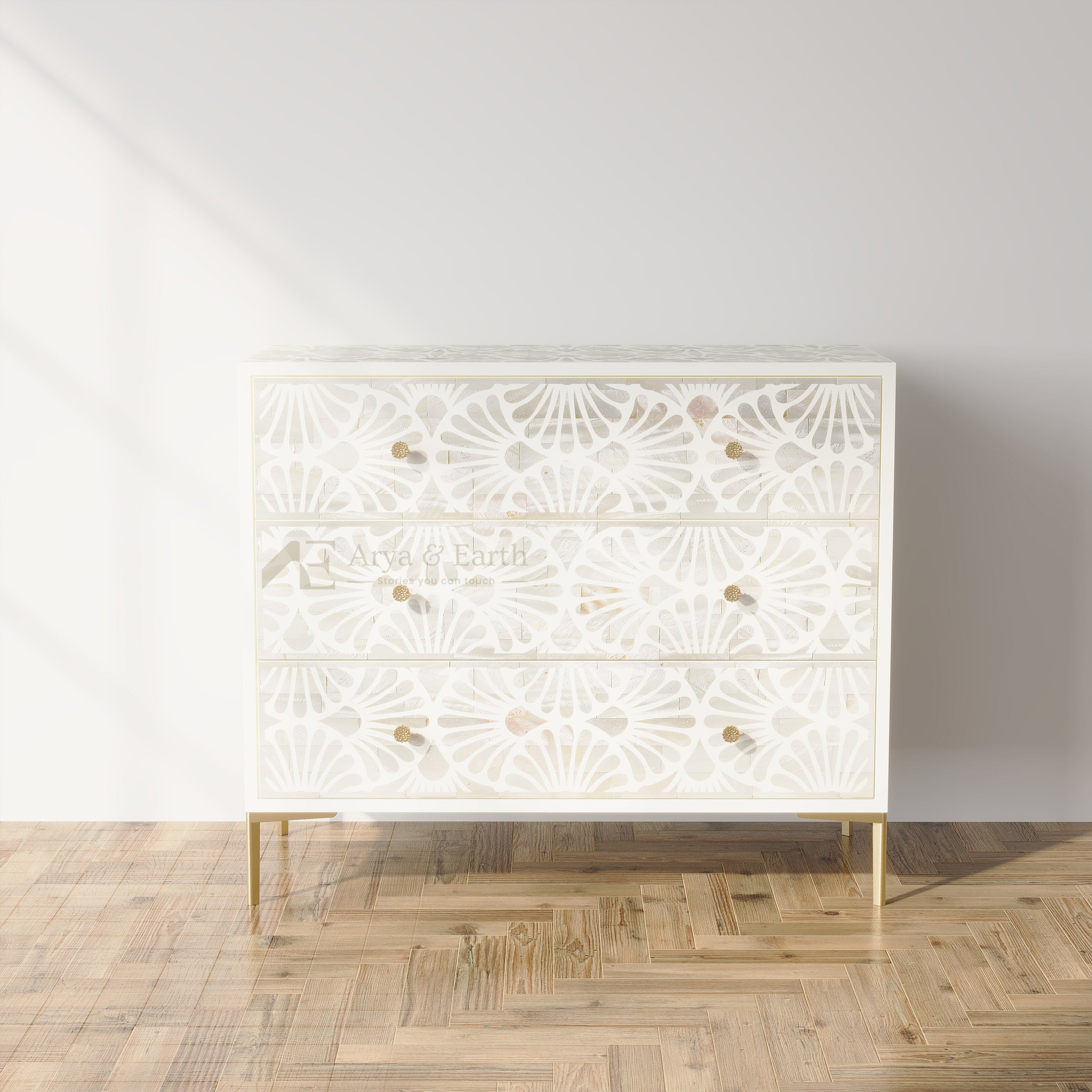 A three-drawer chest of drawers with a white carved geometric floral pattern and small gold knobs, standing on slim gold legs against a light gray wall and herringbone wood floor.
