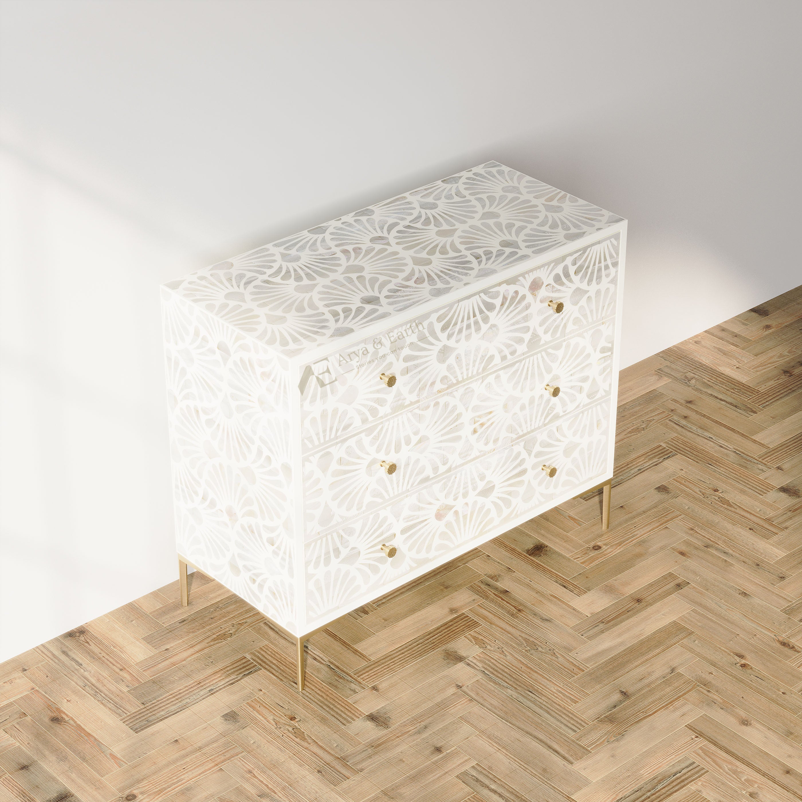 A white three-drawer chest of drawers with an intricate laser-cut fan pattern and gold legs, shown in a styled room setting on a light wood herringbone floor against a white wall.