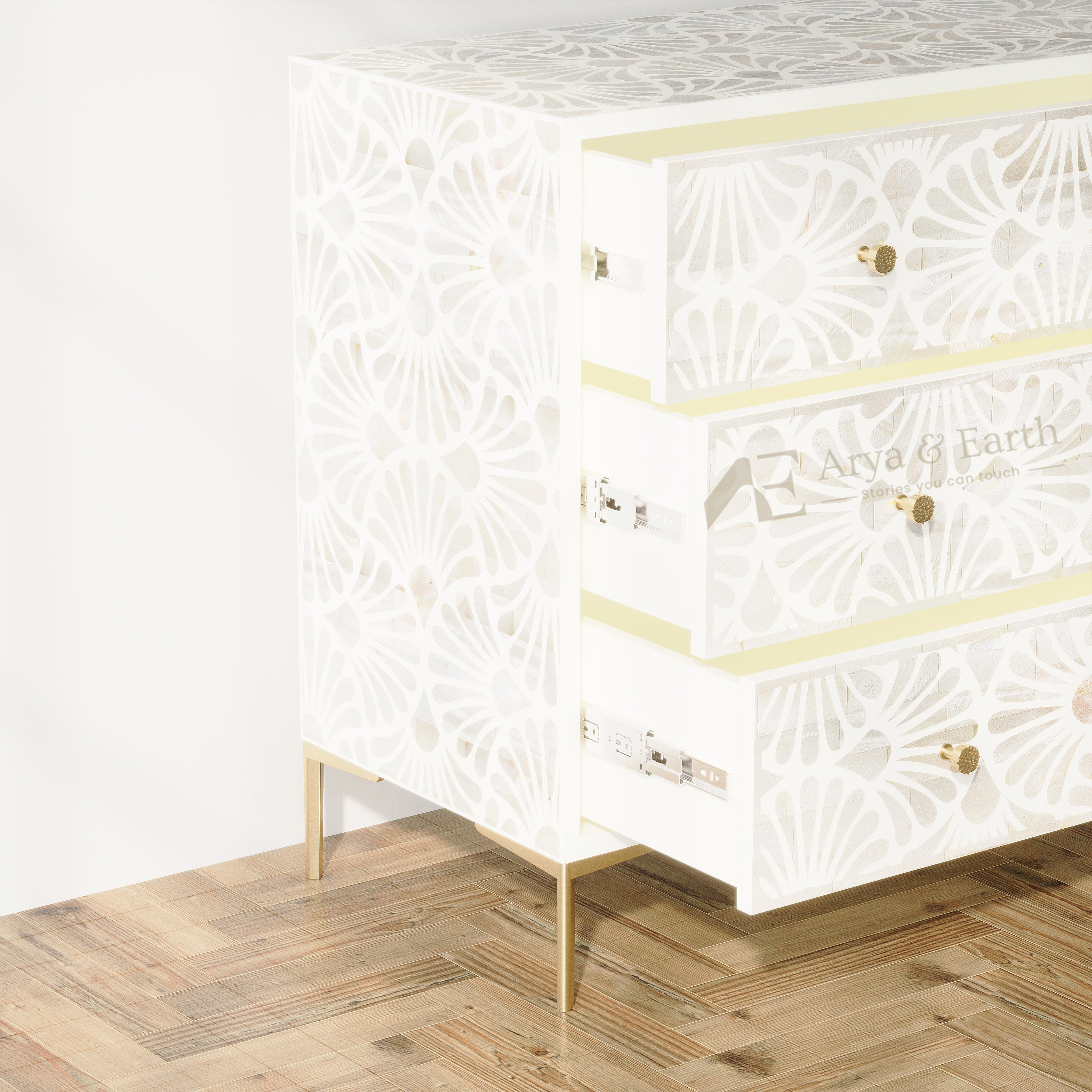 A three-drawer chest of drawers with a white laser-cut floral pattern, pale yellow trim, and gold legs, shown on a wooden parquet floor against a white wall. The brand name 'Arya & Earth' is visible on the drawer front.