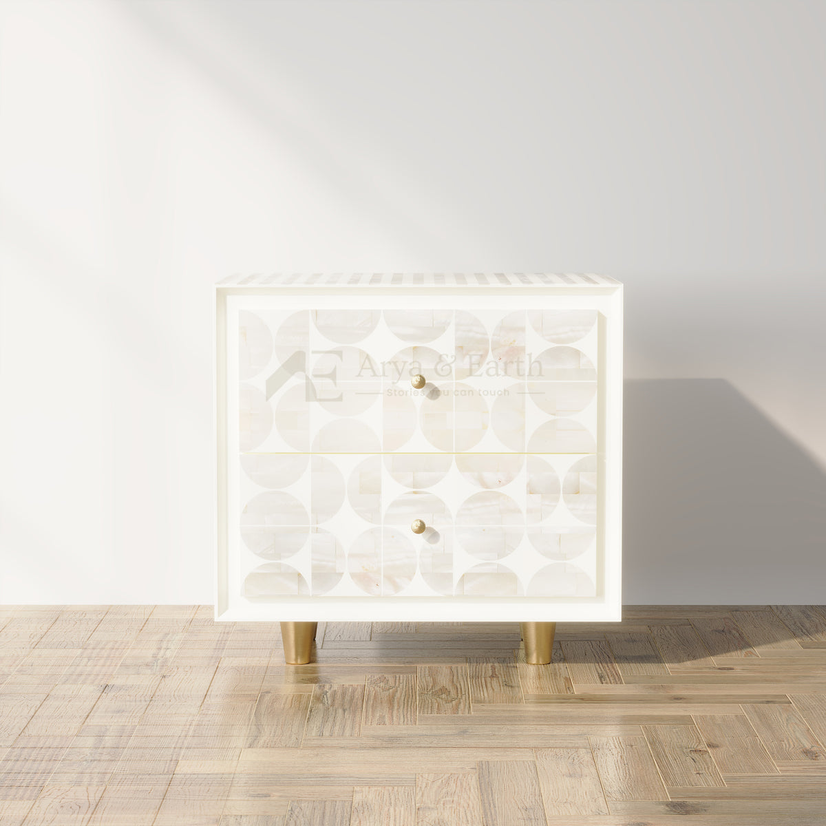 A modern two-drawer chest of drawers with a white frame and back panels featuring a circular shell pattern, supported by gold-toned legs, set against a minimalist white wall and light wood parquet floor.