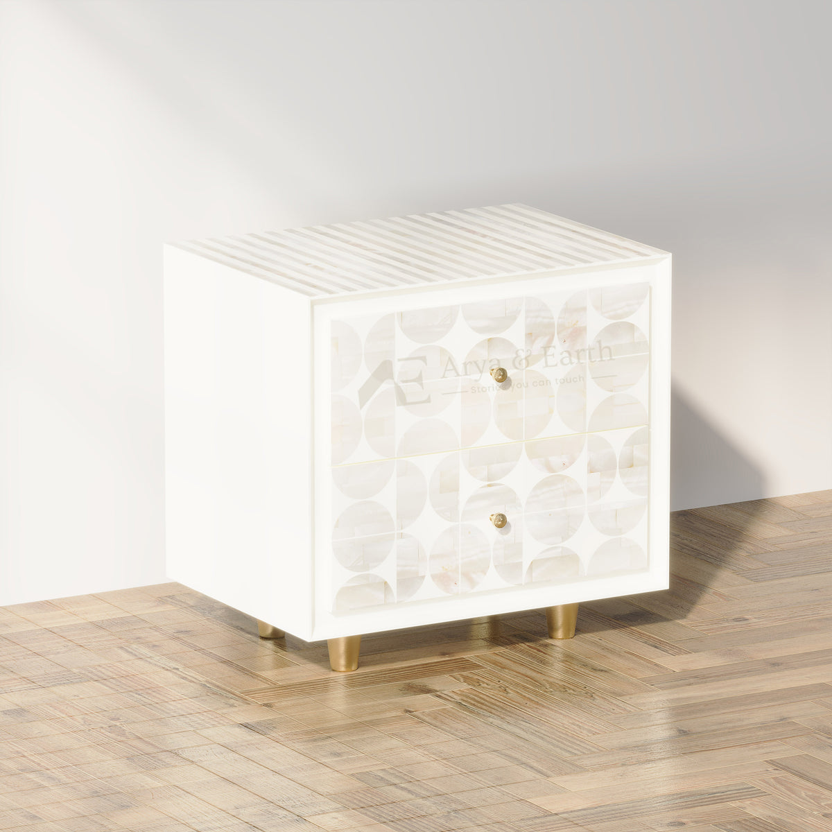 A white wooden nightstand with a mother-of-pearl patterned front, brass legs, and two small brass knobs, set on a light wood herringbone floor against a white wall.