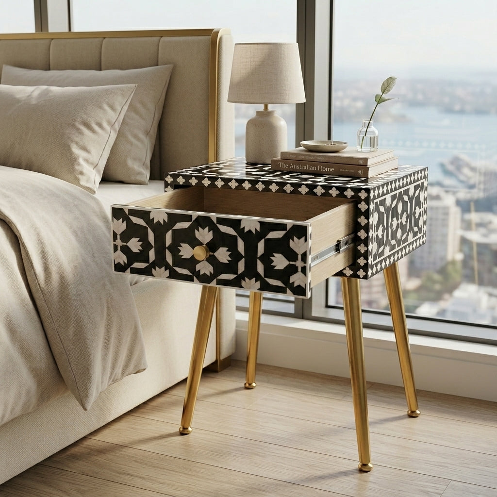 A modern bedside table with a black and white geometric bone-inlay pattern and gold metal legs, styled next to a bed in a bright bedroom with a window view.
