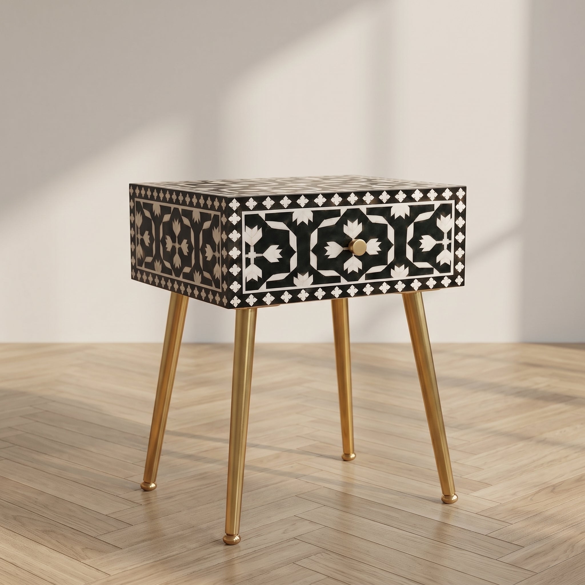 A rectangular side table with a black and white geometric bone-inlay pattern on the drawer front and top, supported by four tapered gold metal legs, set against a neutral beige wall and light wood herringbone floor.
