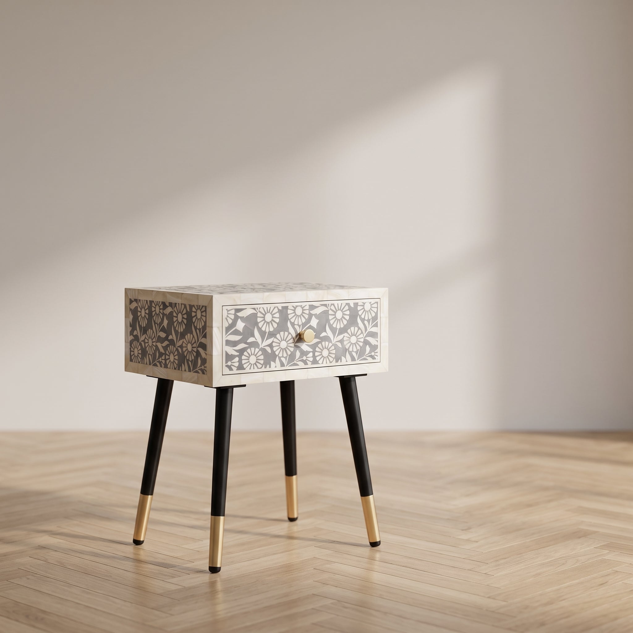 A small rectangular side table with a single drawer, featuring a white and gray floral bone-inlay pattern on the front and side panels, black tapered legs, and gold-toned metal caps, set on a light wood herringbone floor against a neutral beige wall with soft window light.