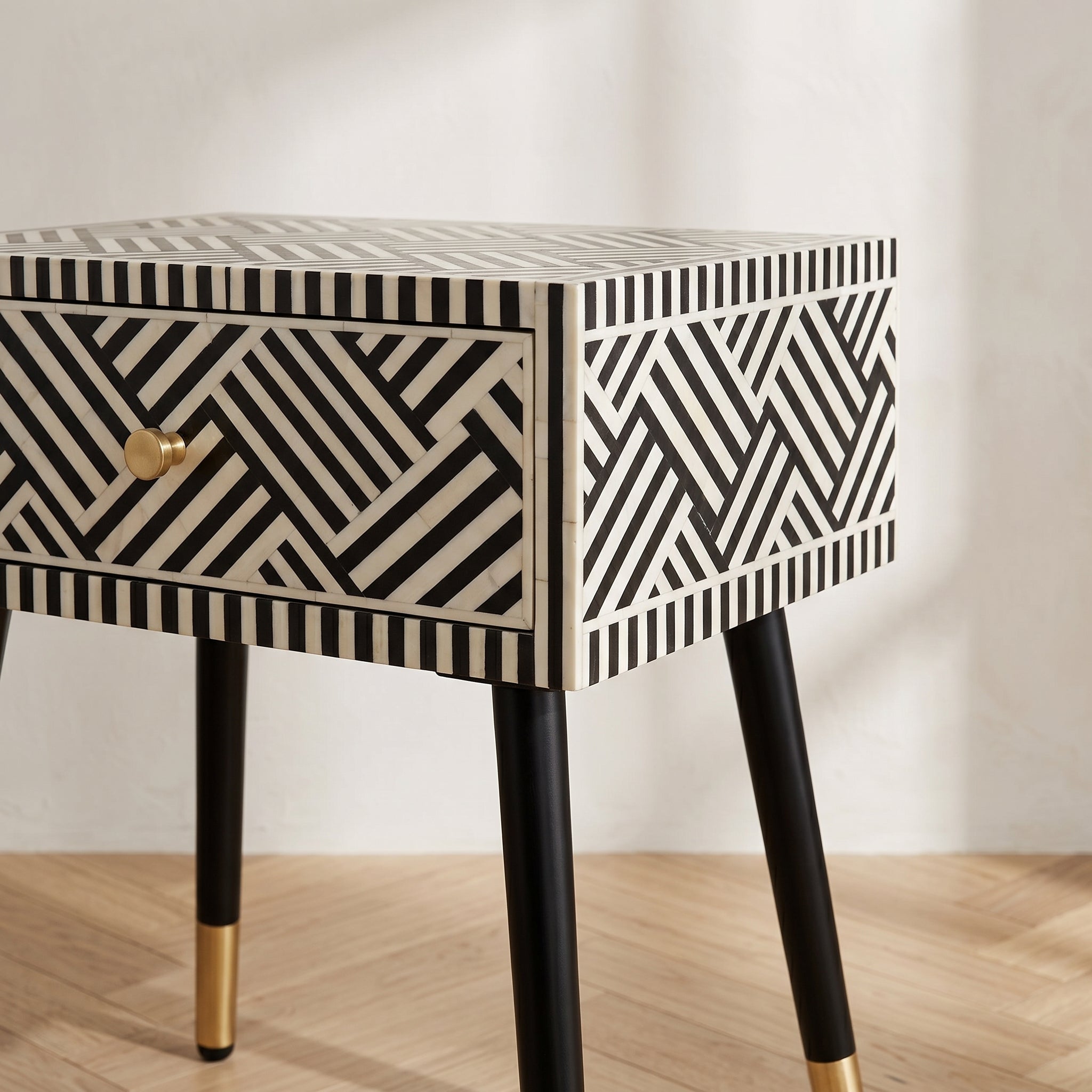 A modern side table with a black and cream geometric bone-inlay pattern, featuring a single drawer with a gold knob and black legs with gold caps, set against a neutral background.