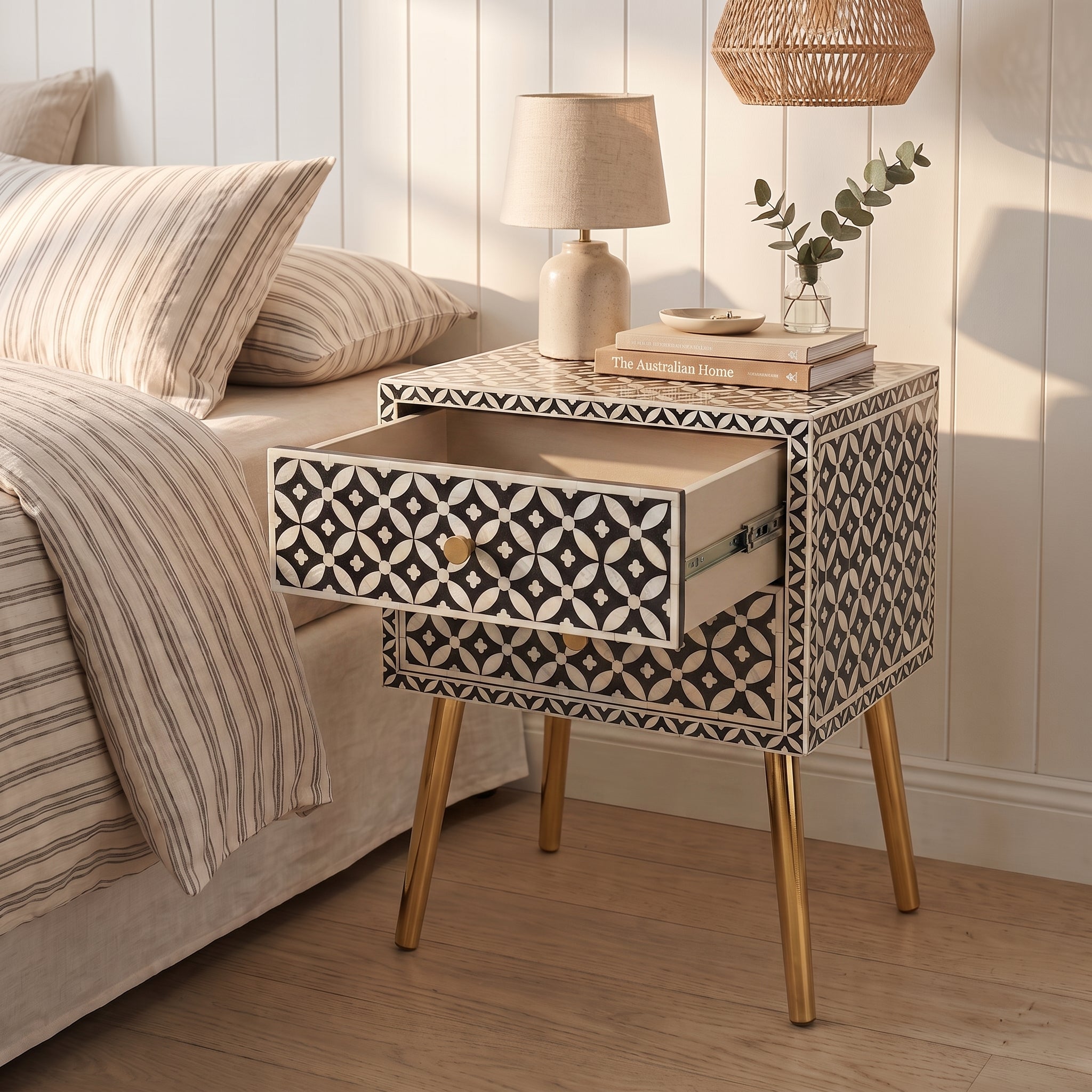 A mid-century modern bone-inset nightstand with geometric black and cream patterns and gold legs, styled next to a bed in a bright bedroom.