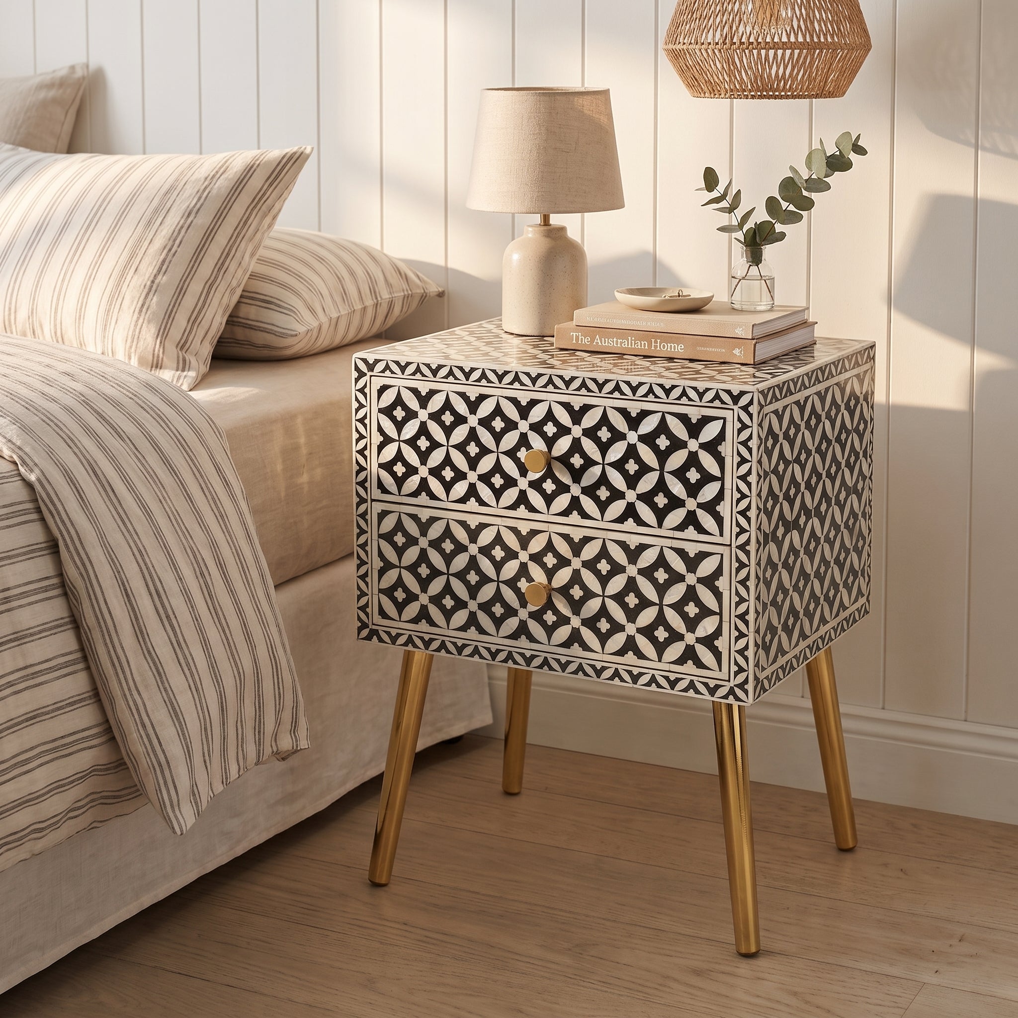 A two-drawer bedside table with a black and white geometric bone-inlay pattern and gold-tone legs, styled next to a bed in a bright bedroom.